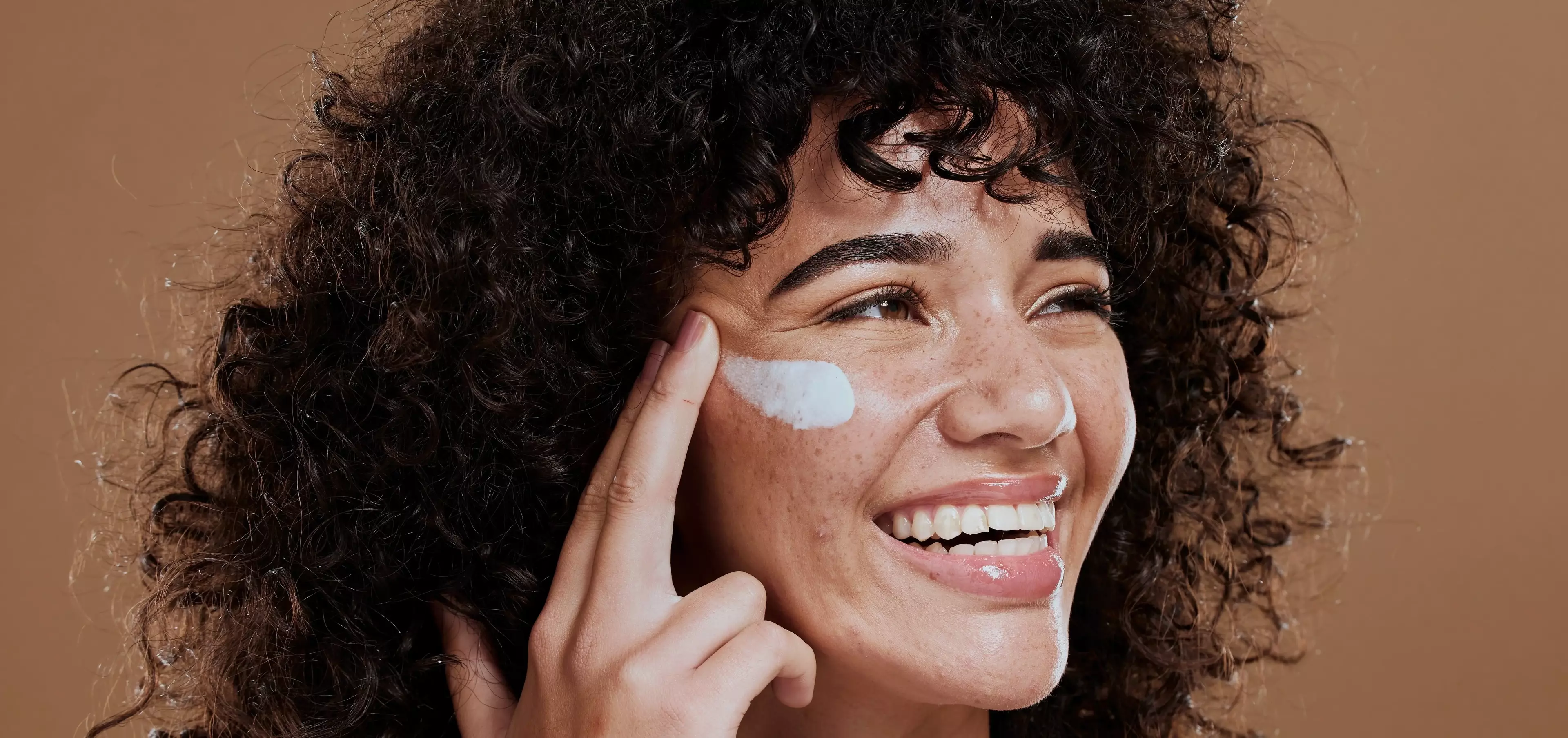 Smiling Woman putting cream on her face with a beige background.